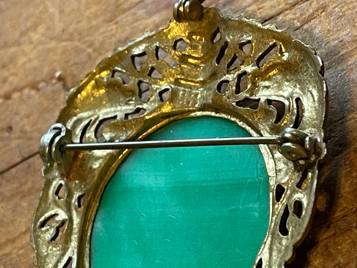 Close-up of a vintage filigree brooch with a bright turquoise green oval center and an ornate gold-toned metal border, showing the pin clasp on a rustic wooden surface.