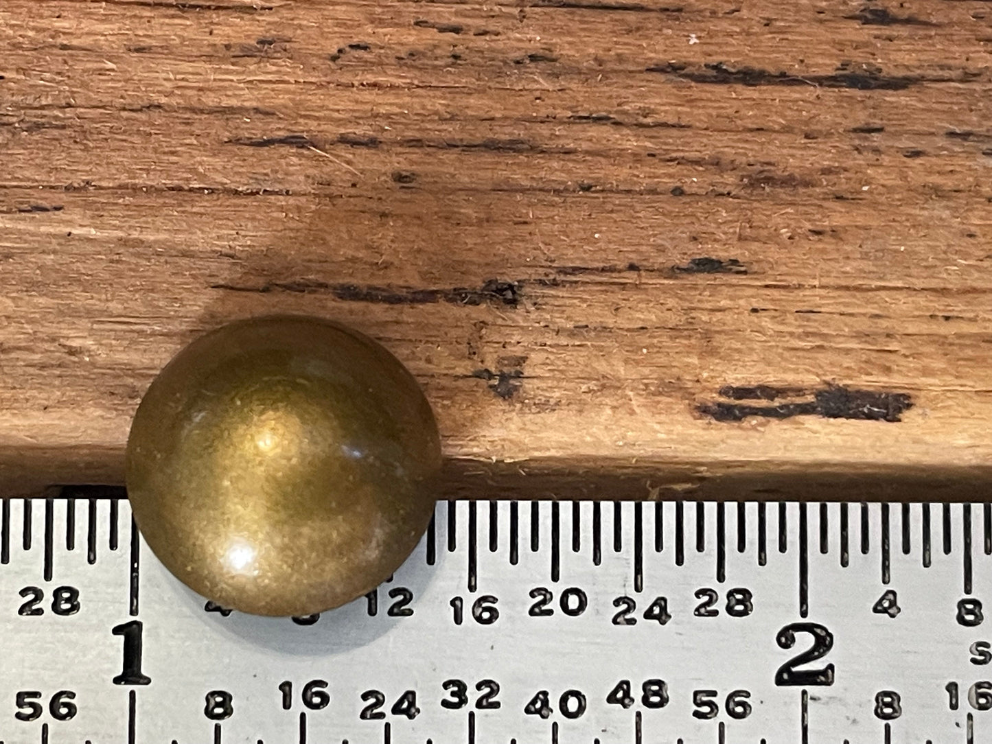 A round, metallic gold-colored upholstery tack seated between a wooden surface and a metal ruler for scale.