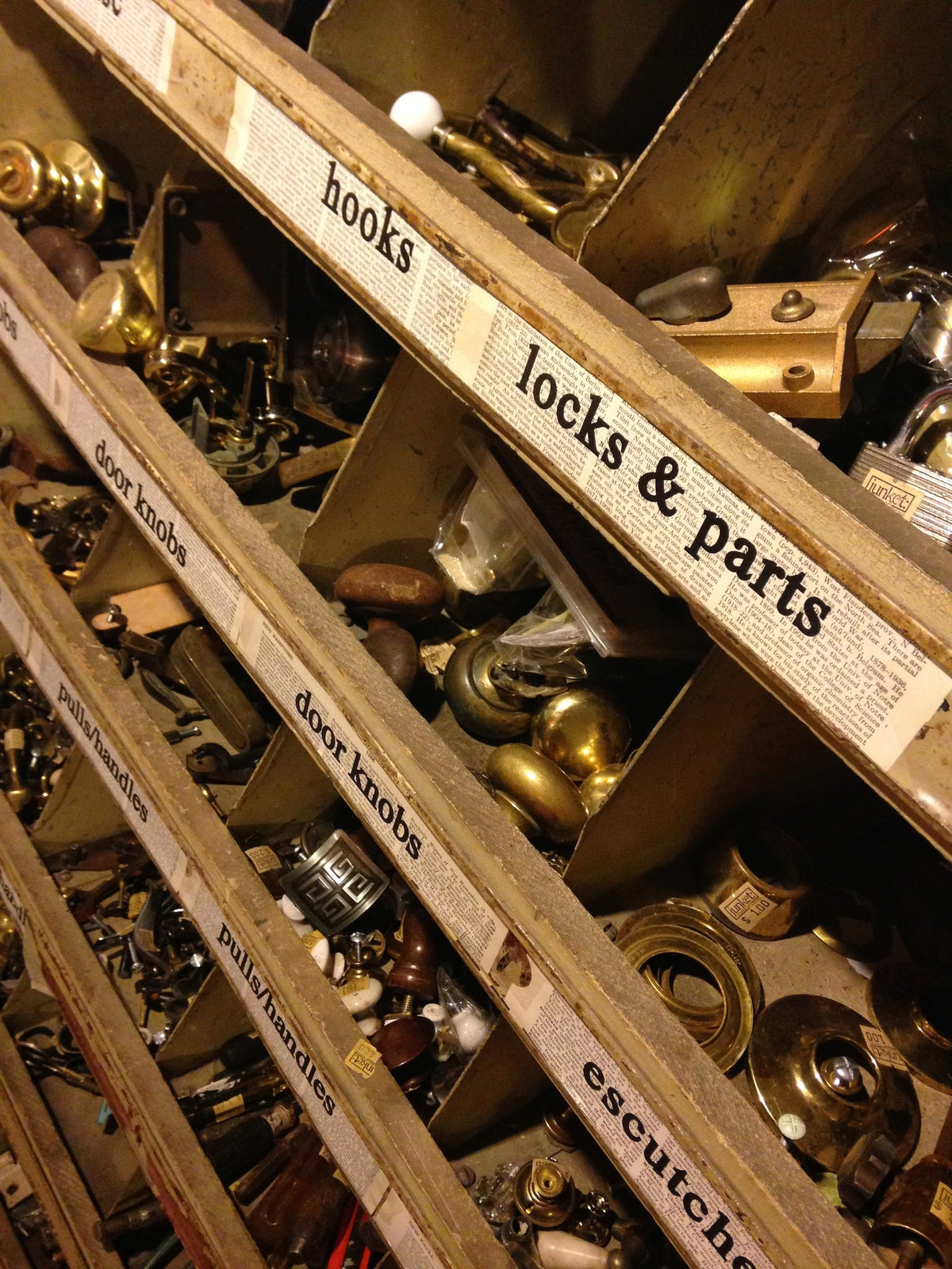 Antique Industrial Parts Bin - a repurposed retail fixture in the old shop basement - holding various types of sorted hardware and labeled with hardware part names printed on old book pages ('locks & parts, hooks, door knobs, escutcheons, pulls/handles).
