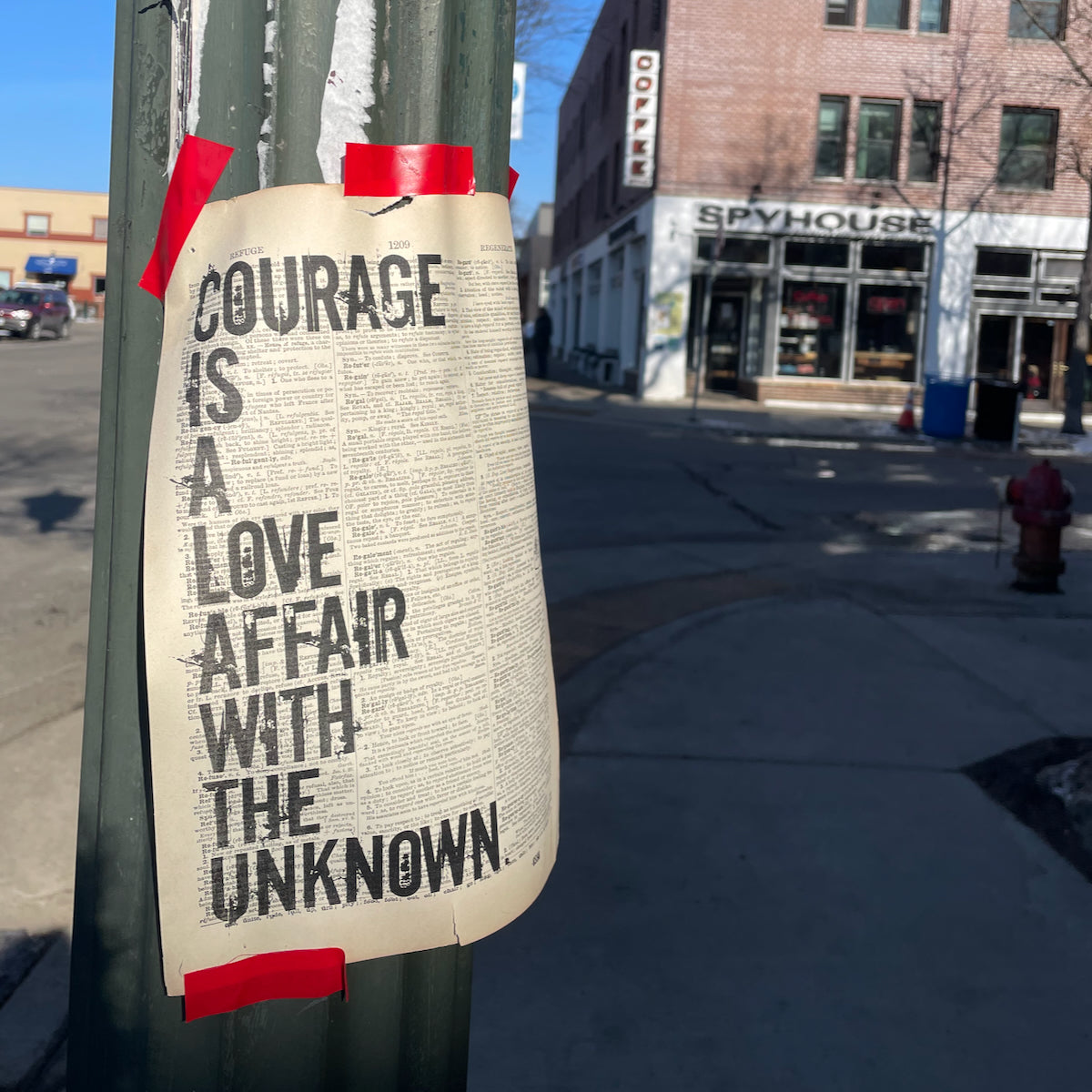 Quotation 'courage is a love affair with the unknown' printed on a dictionary page, and taped to a lightpost with 'Spyhouse' coffee shop behind it.