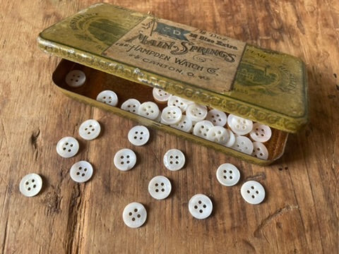 White round buttons scattered on a rustic wooden surface next to an open vintage metal tin filled with more buttons.