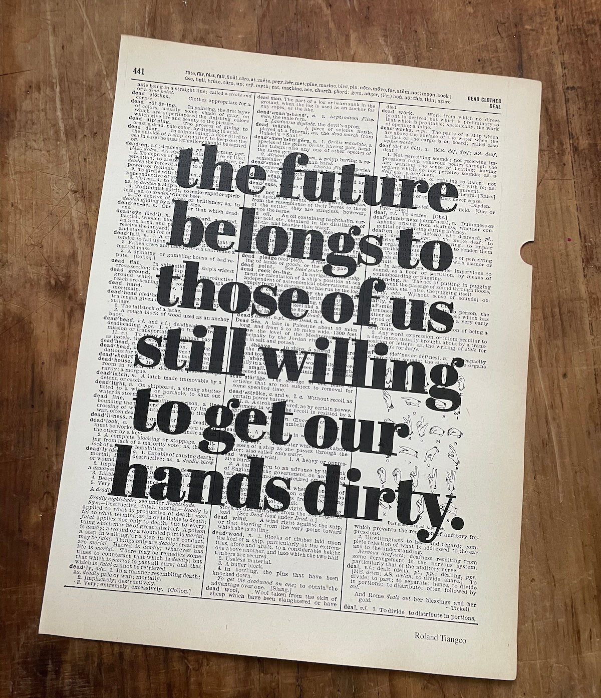 Dictionary page with overprinted motivational quote on a wooden surface: "the future belongs to those of us still willing to get our hands dirty."
