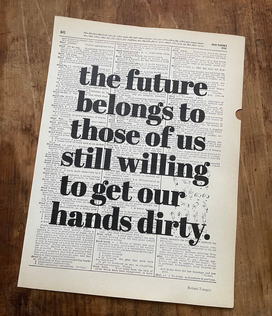 Dictionary page with overprinted motivational quote on a wooden surface: "the future belongs to those of us still willing to get our hands dirty."