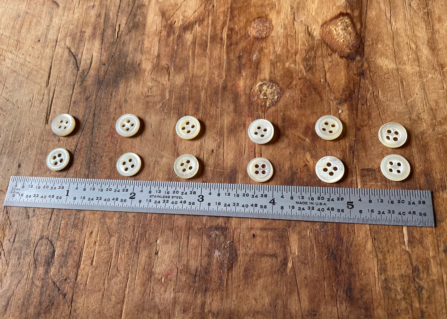 Twelve small cream-colored four-button sewing buttons arranged vertically next to a metal ruler on a rustic wooden surface.