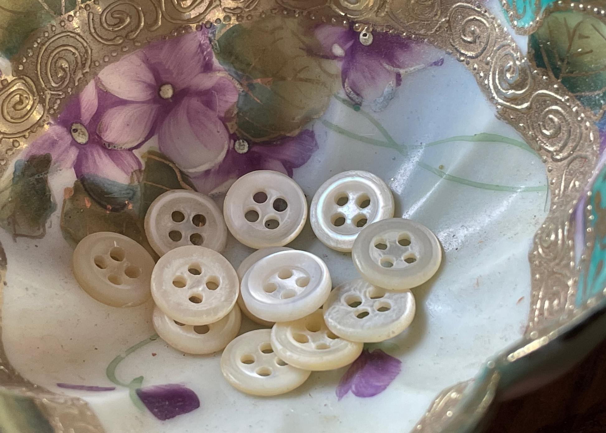 A group of round, four-hole buttons made of mother-of-pearl, arranged inside a decorative ceramic dish with purple flowers and a gold-toned rim.