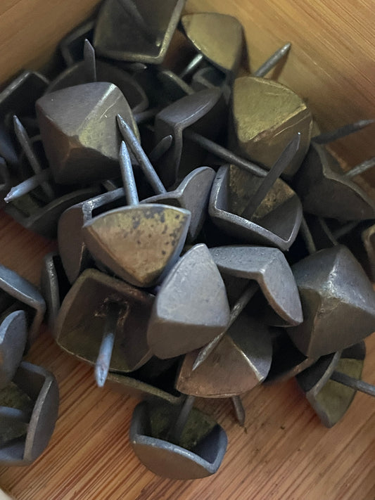 A pile of geometric brass upholstery tacks with pyramid shaped heads and  steel shanks, resting on a light brown wooden surface.