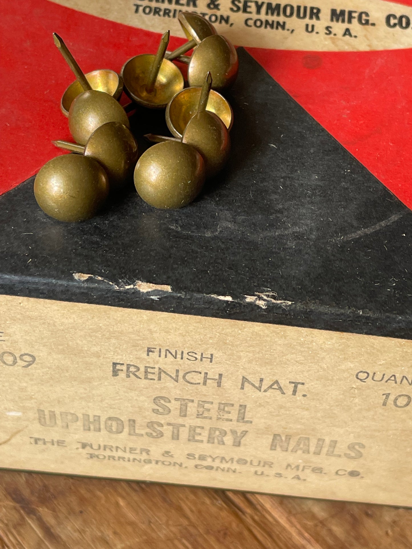 A close-up of steel upholstery nails resting on a vintage paper box labeled 'French Nat. Finish Steel Upholstery Nails' on a wooden surface.