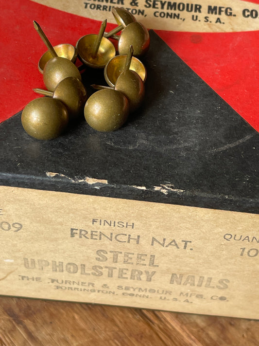 A close-up of steel upholstery nails resting on a vintage paper box labeled 'French Nat. Finish Steel Upholstery Nails' on a wooden surface.