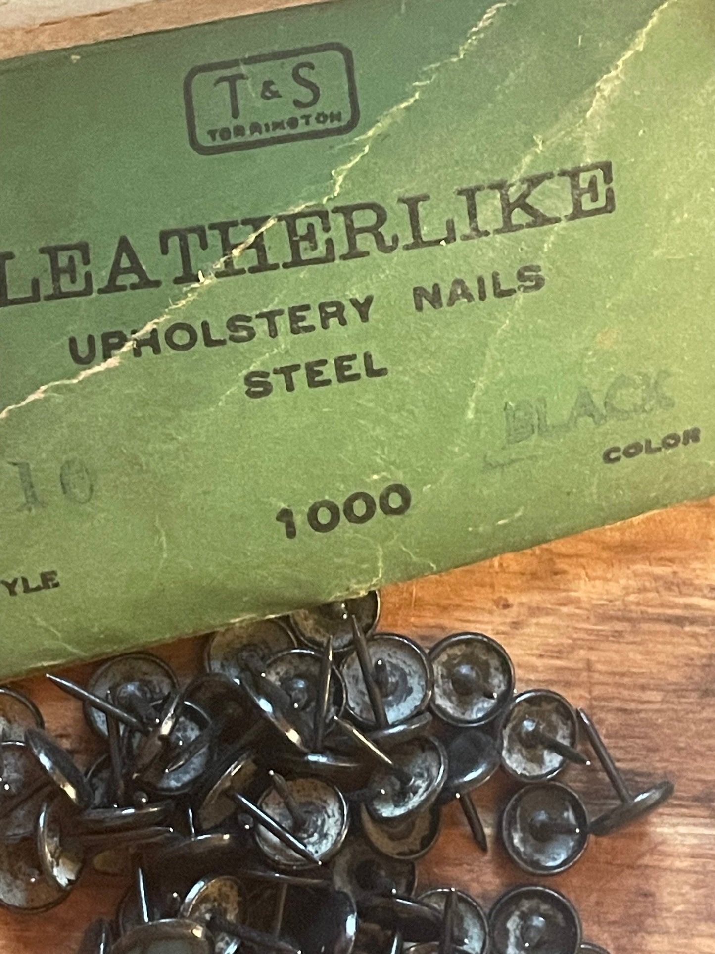 A close-up of a green T&S Leatherlike Upholstery Nails 1000 Steel product box next to a pile of black metal upholstery nails on a wooden surface.