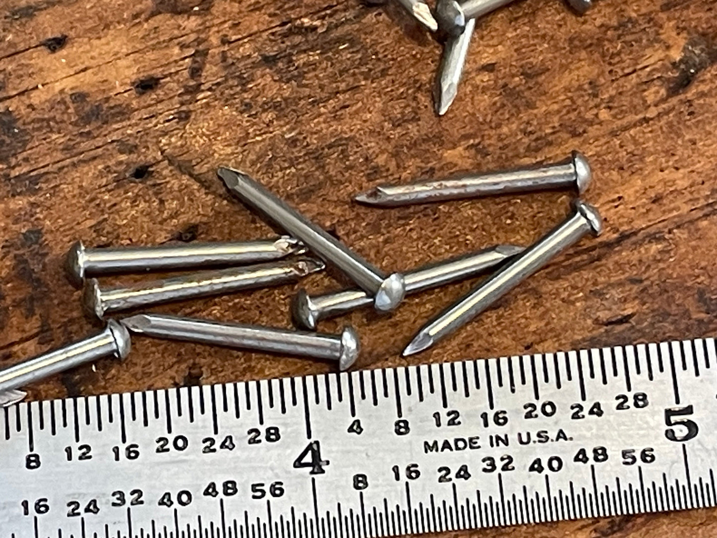 Several silver metal nails scattered on a rustic wooden surface next to a metal ruler for scale.