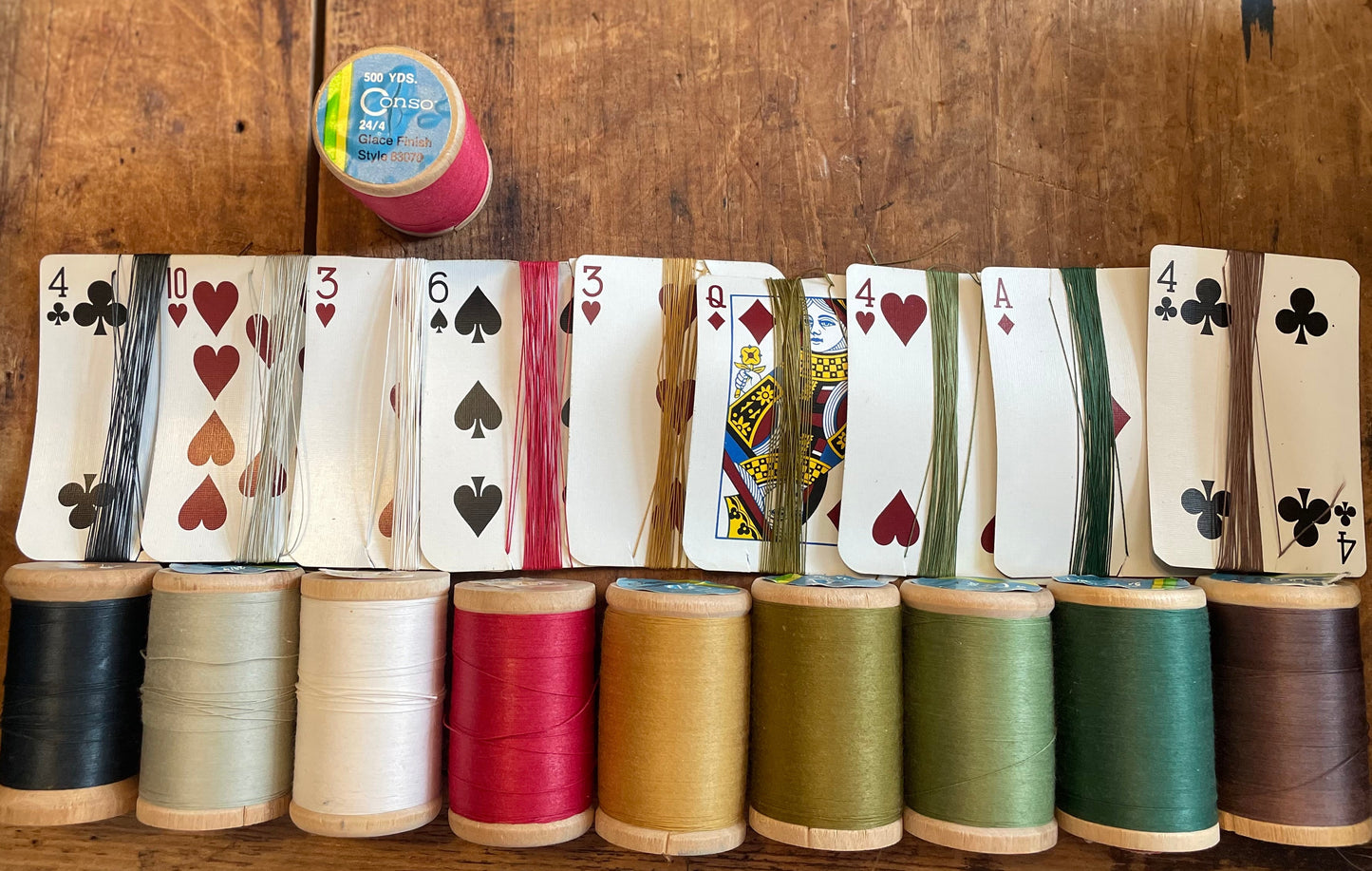 A set of nine spools of thread in various colors, each layered with a corresponding playing card wrapped in that spool's thread and displayed on a rustic wooden table.