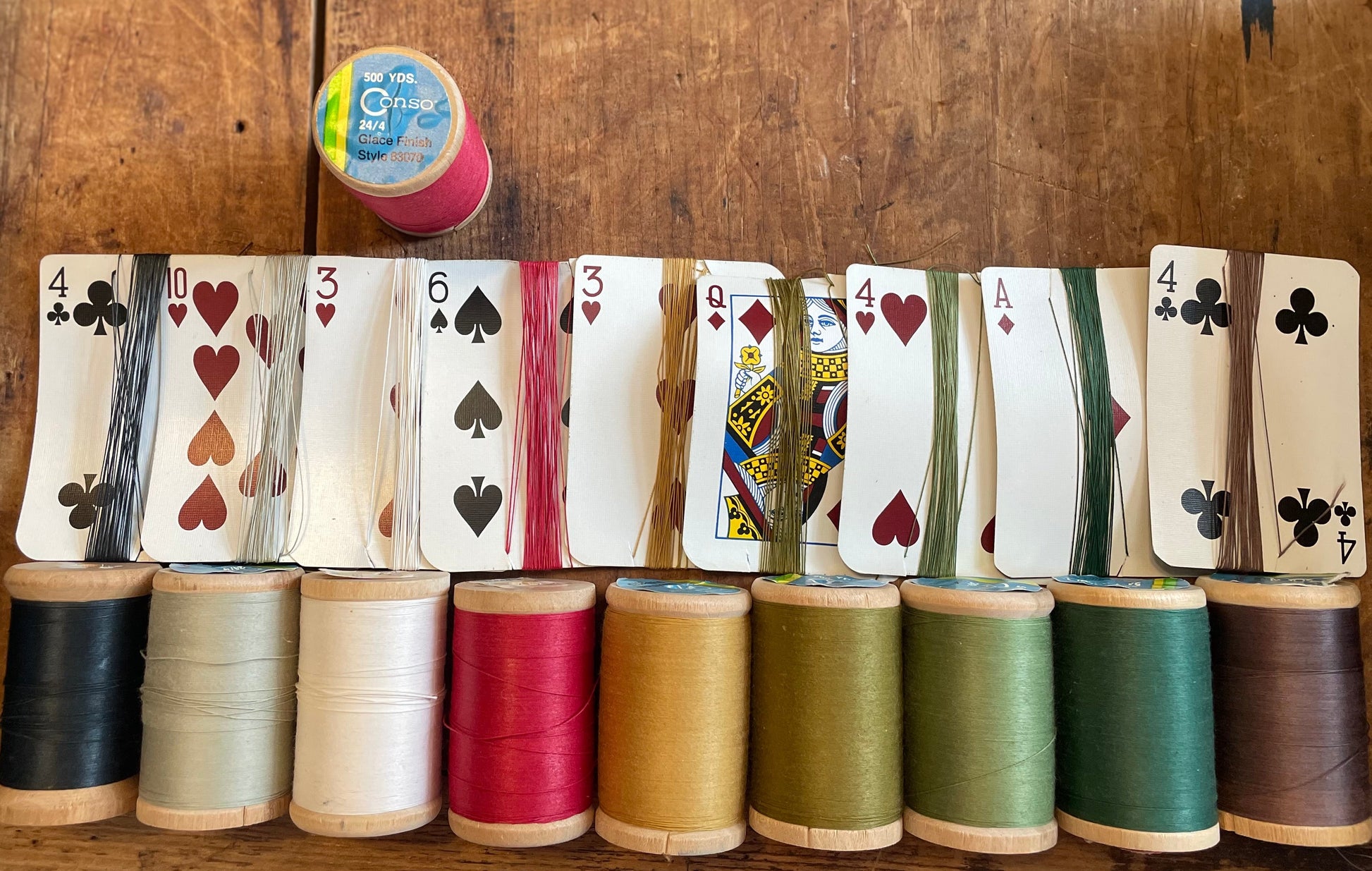 A set of nine spools of thread in various colors, each layered with a corresponding playing card wrapped in that spool's thread and displayed on a rustic wooden table.