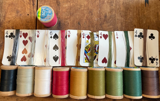A set of nine spools of thread in various colors, each layered with a corresponding playing card wrapped in that spool's thread and displayed on a rustic wooden table.