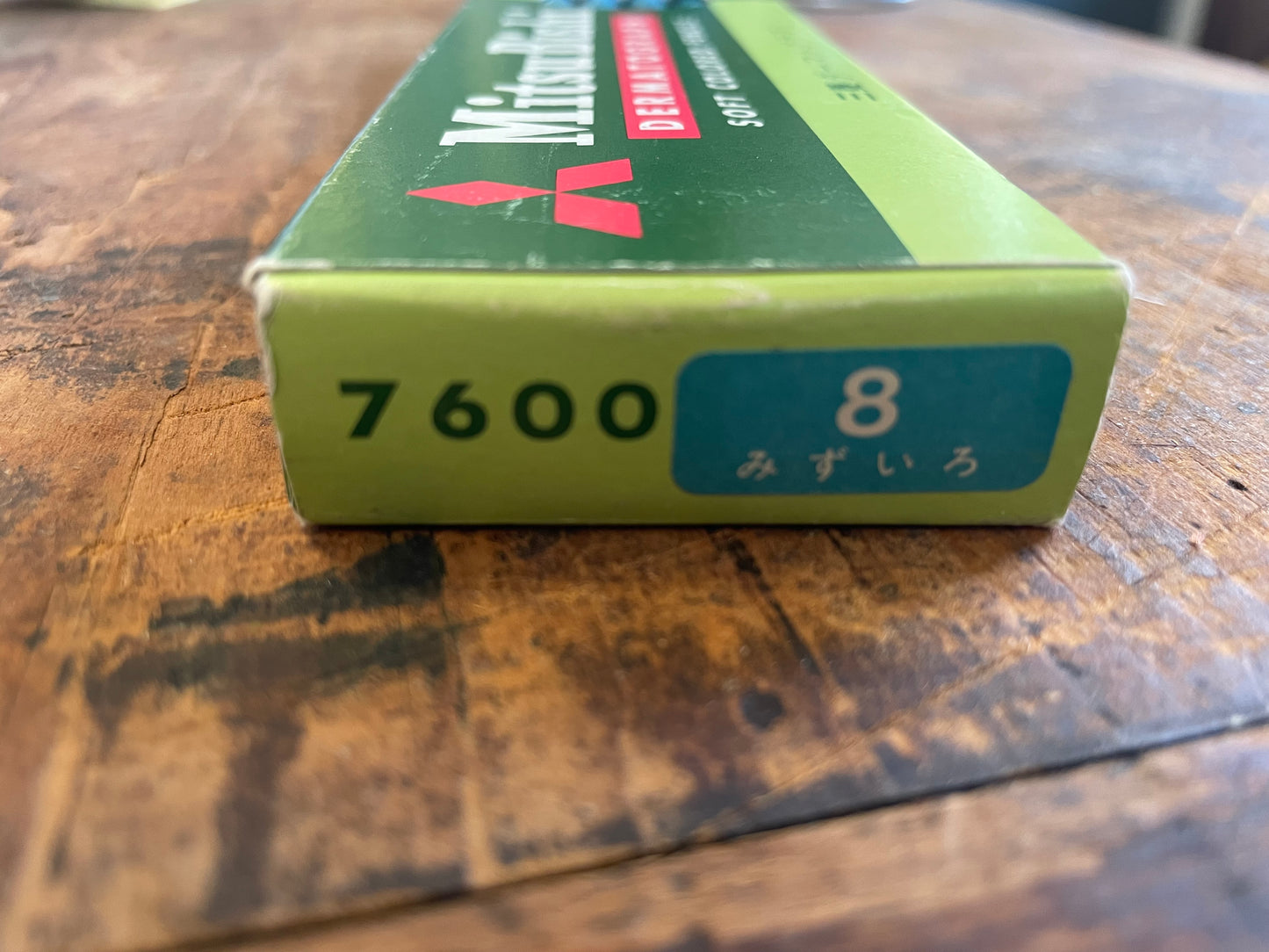 A close-up of a green and light green box of Mitsubishi 7600 8 Dermatograph pencils, resting on a rustic wooden surface.