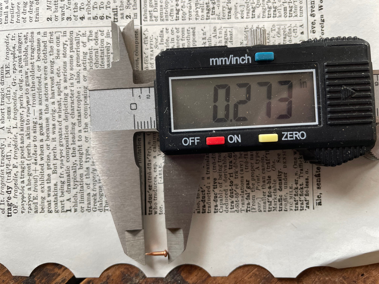 A digital caliper displaying 0.273 inches is shown measuring the length of a small escutcheon pin over a printed book page background.