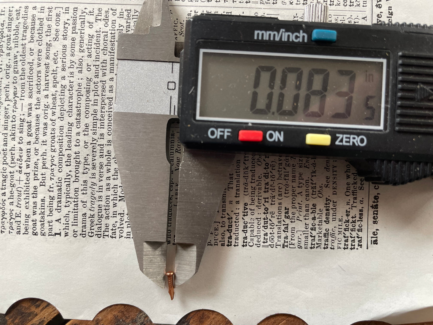 A digital caliper measuring the head diameter of a small escutcheon pin on top of printed text, with the caliper display reading 0.083 inches.