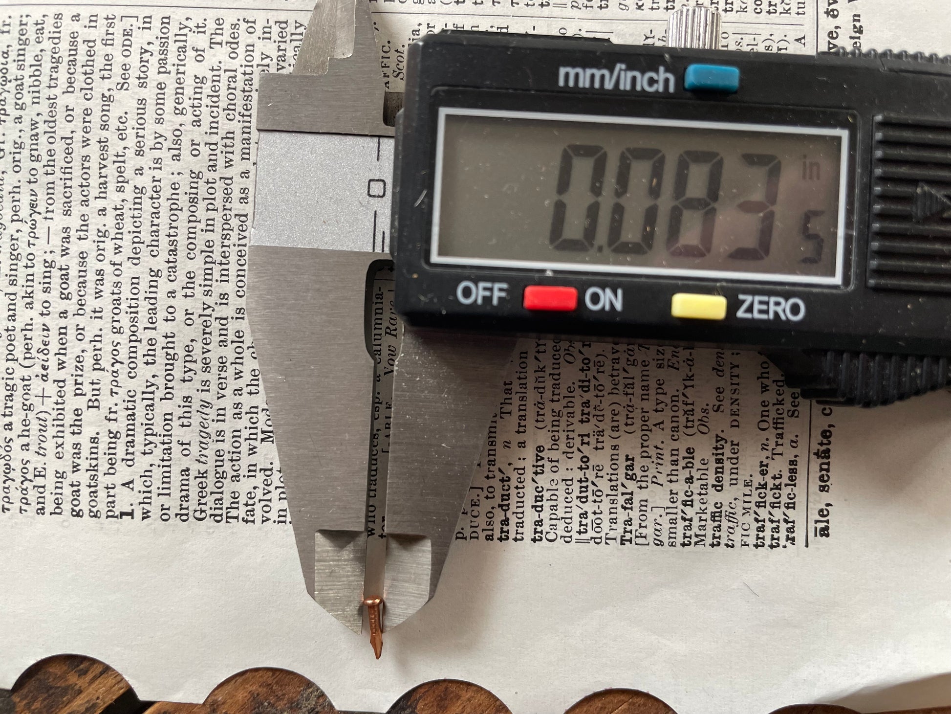 A digital caliper measuring the head diameter of a small escutcheon pin on top of printed text, with the caliper display reading 0.083 inches.