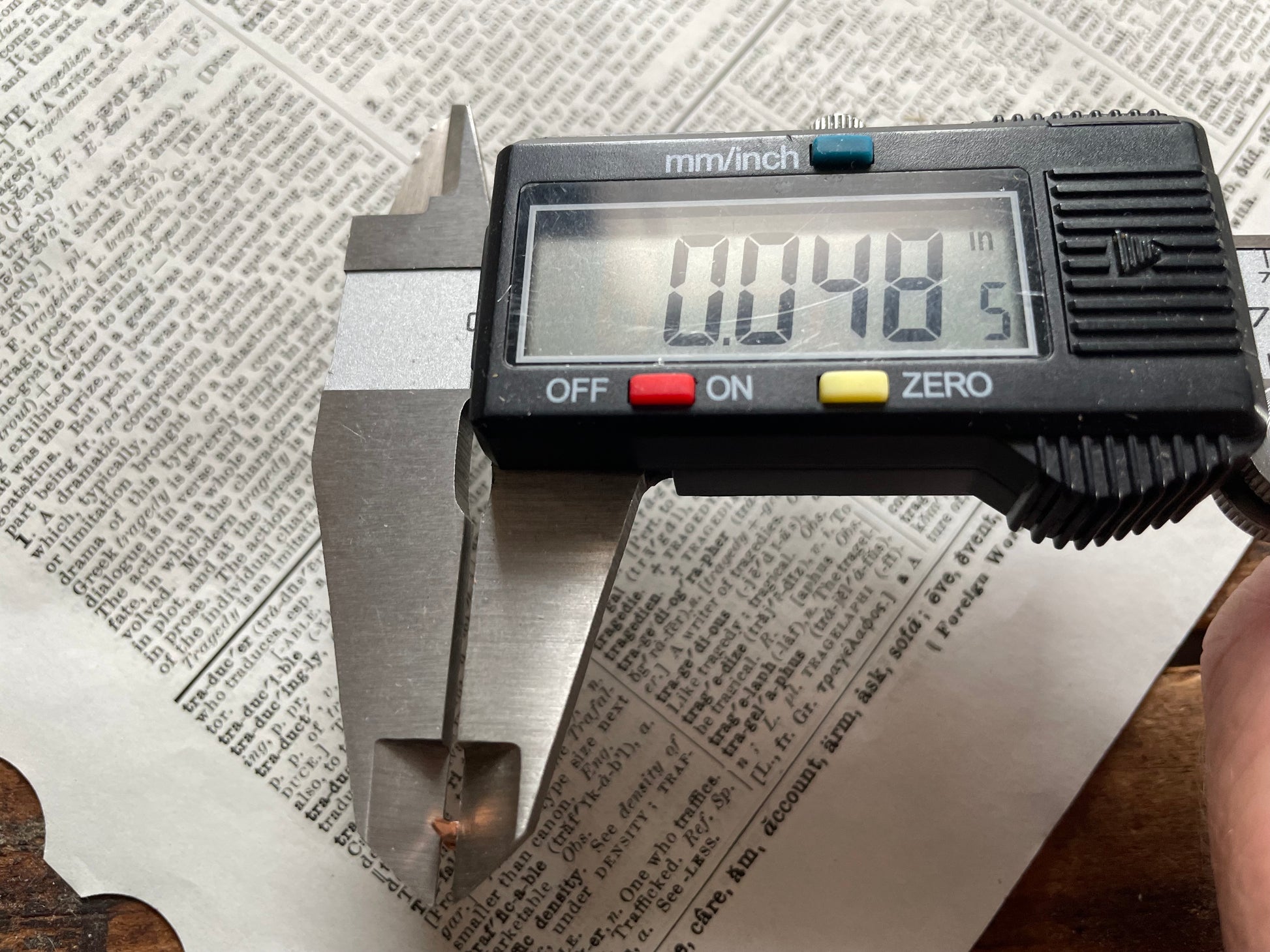 A digital caliper measuring tool measures the pin diameter of a small escutcheon pin (caliper reads 0.48 inches on its LCD screen), positioned over a printed paper background.