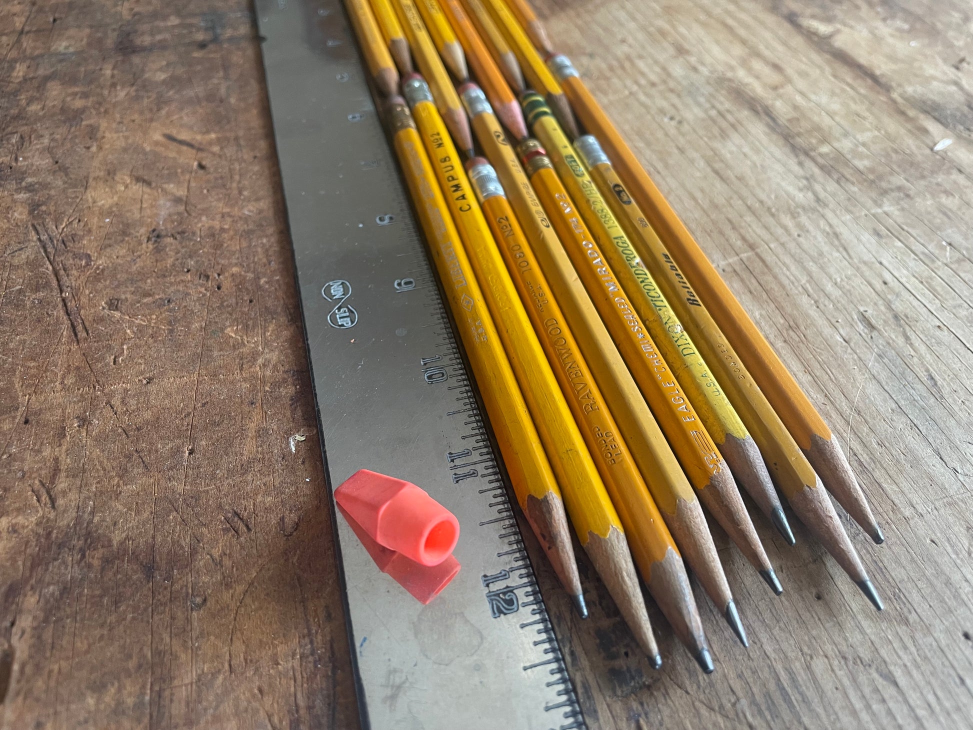 A set of yellow wooden pencils with silver ferrules and gray graphite tips arranged next to a metal ruler on a worn wooden tabletop. A small orange eraser is also visible.