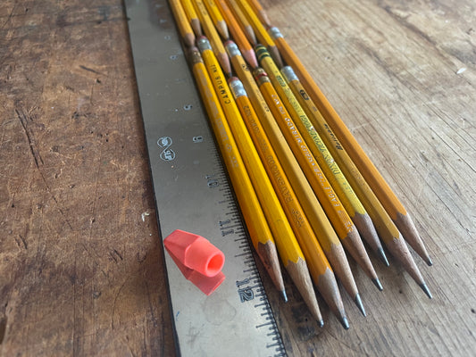 A set of yellow wooden pencils with silver ferrules and gray graphite tips arranged next to a metal ruler on a worn wooden tabletop. A small orange eraser is also visible.