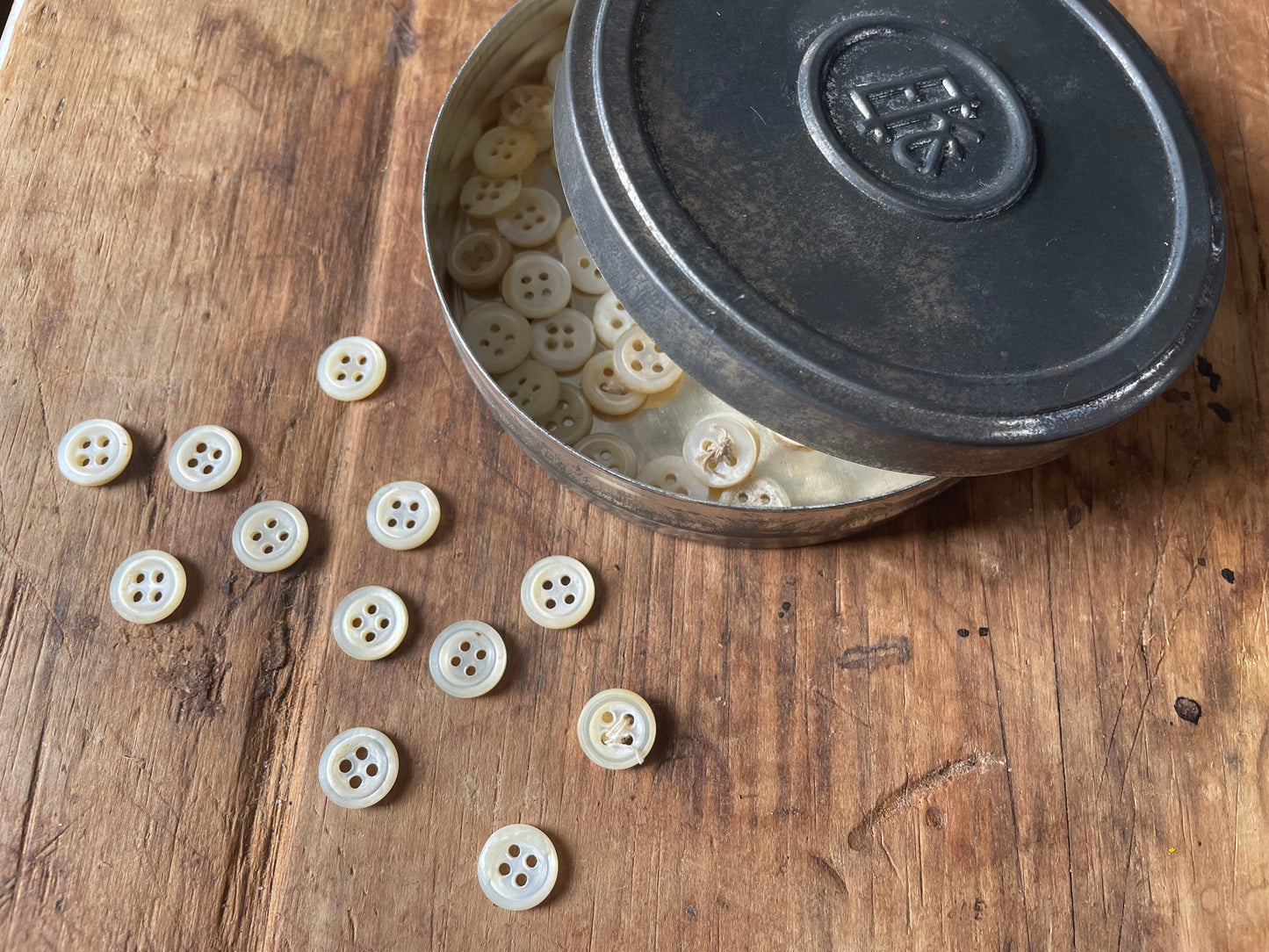 Cream-colored four-hole buttons scattered on a rustic wooden surface next to an open dark metal tin filled with more buttons.