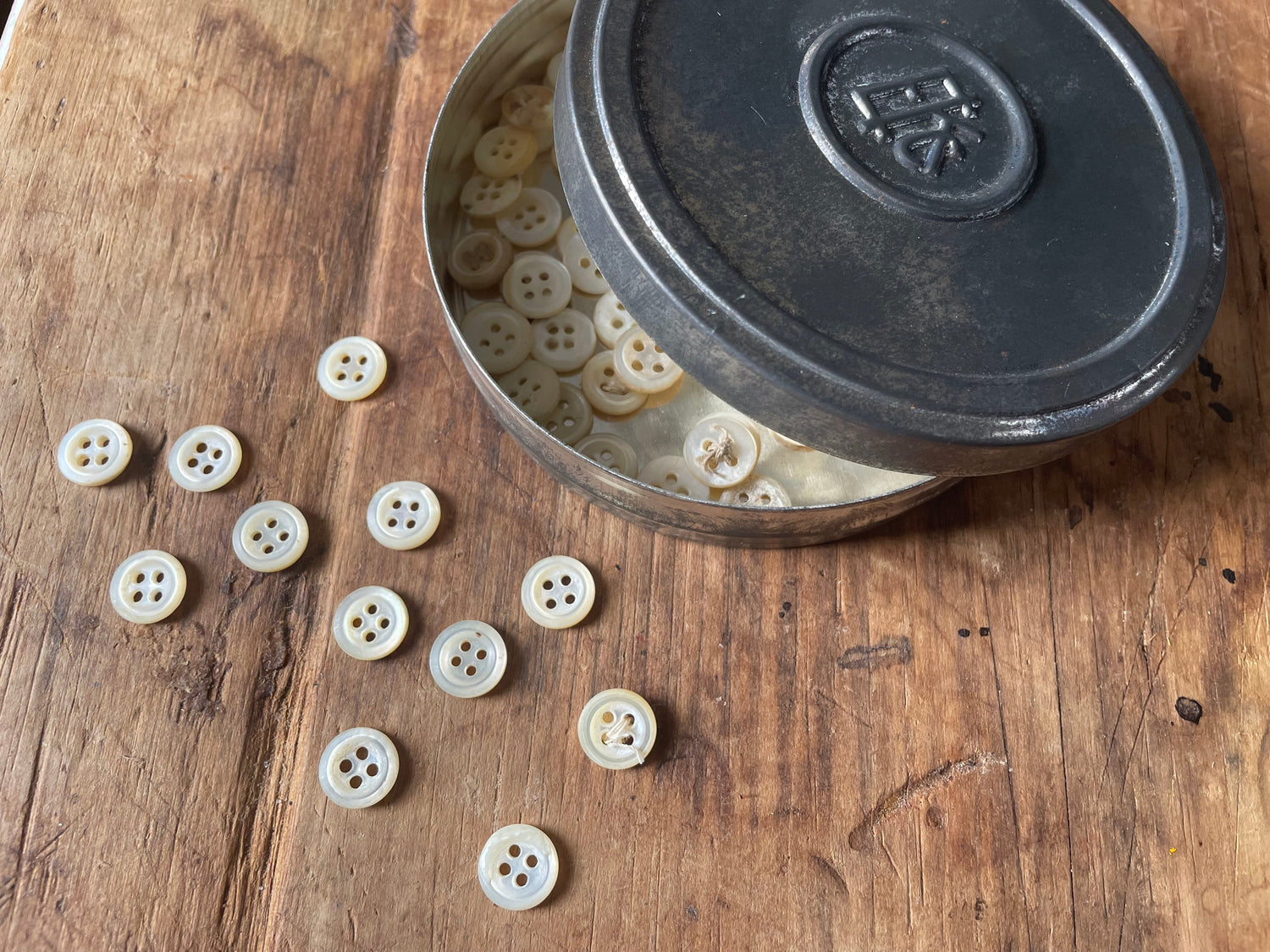 Cream-colored four-hole buttons scattered on a rustic wooden surface next to an open dark metal tin filled with more buttons.