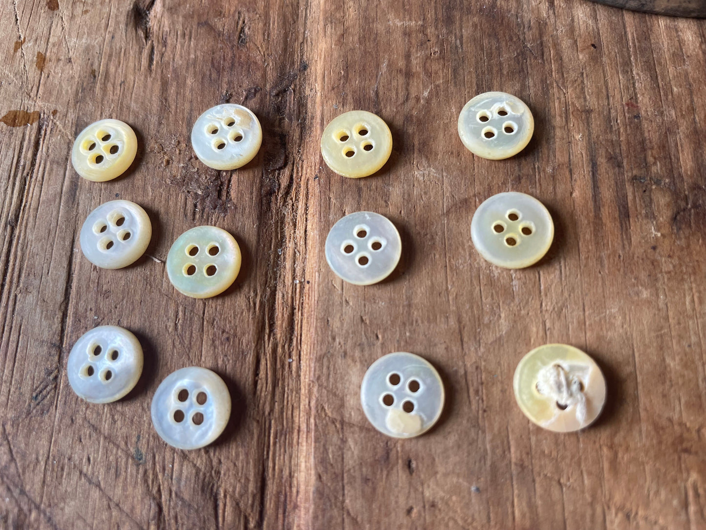 Twelve small round buttons with four holes, made from natural mother-of-pearl, arranged with their back sides facing the camera on a rustic wooden surface. The buttons have iridescent, pearlescent colors ranging from cream and pale yellow to light gray.