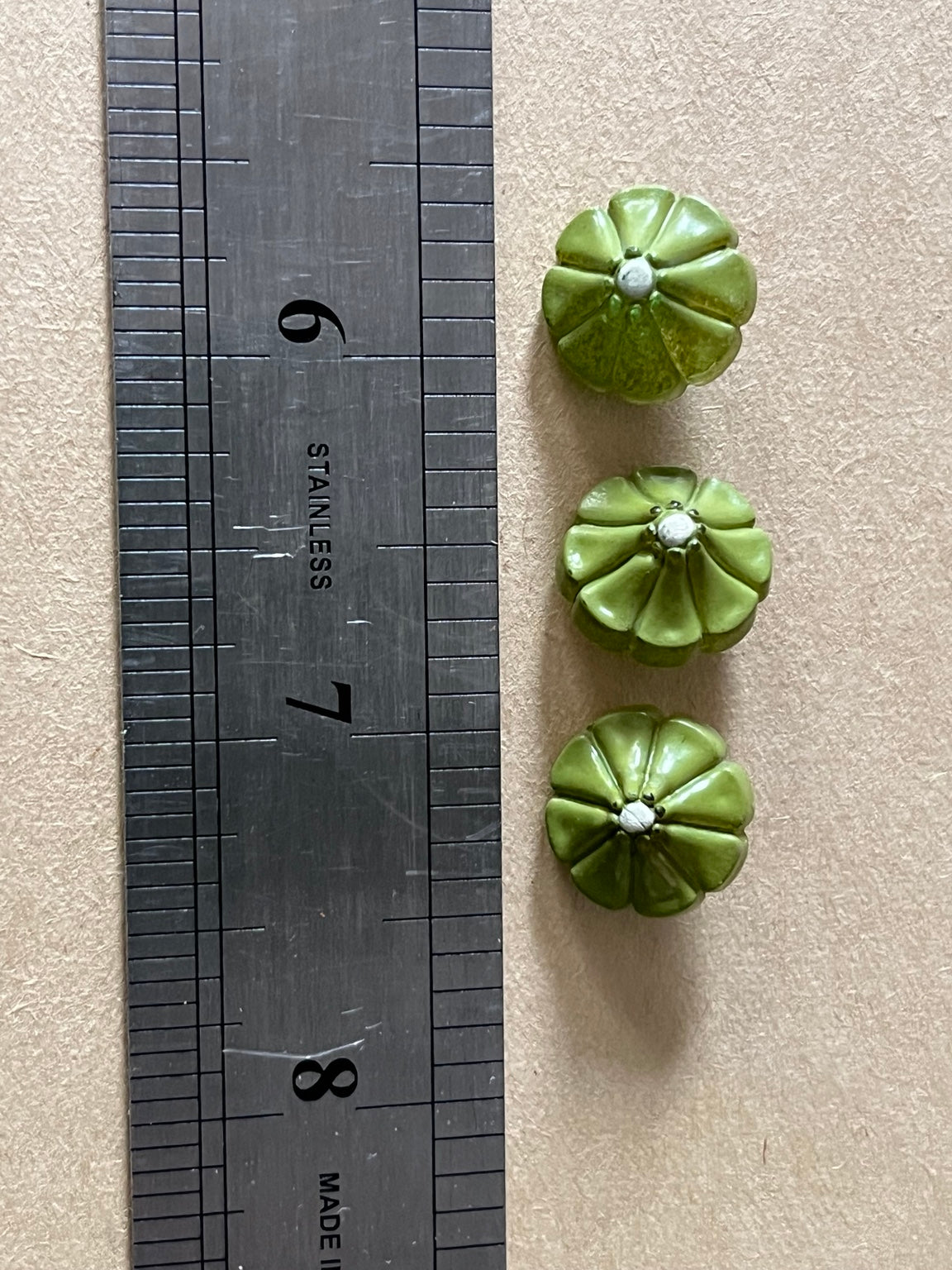 Three olive green flower-shaped buttons beside a metal ruler on beige paper.