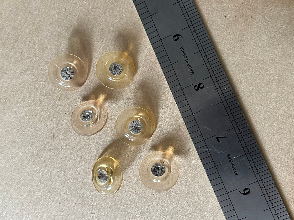 Six translucent pale champagne round buttons with clear faceted centers beside a metal ruler on beige paper.