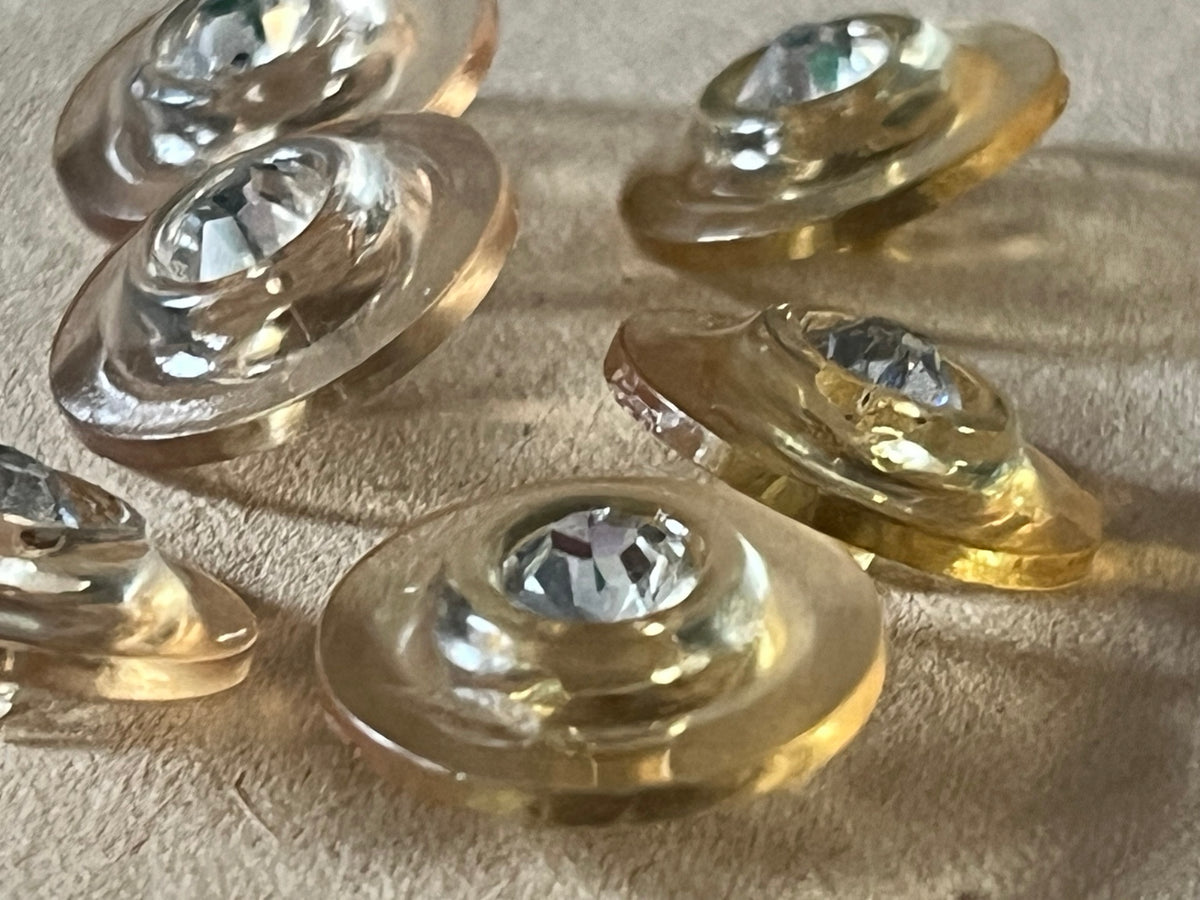 Six translucent pale champagne round buttons with clear faceted centers in an angled close-up on beige paper.
