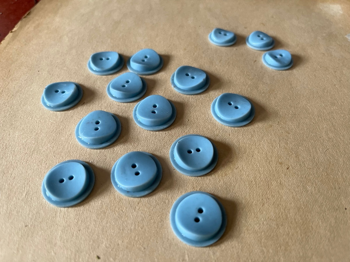 Fifteen powder blue round two-hole buttons in mixed sizes on brown paper.