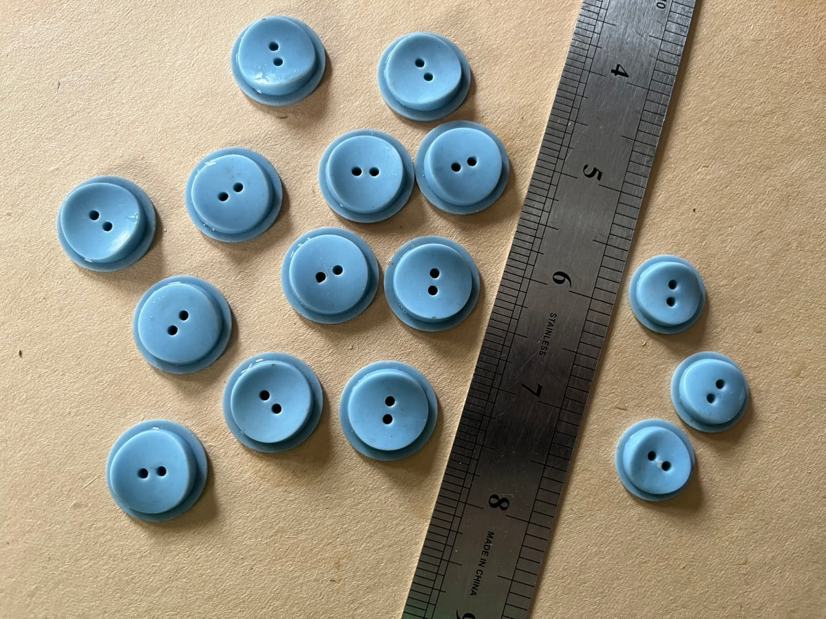 Fifteen powder blue round two-hole buttons in mixed sizes beside a metal ruler on brown paper.