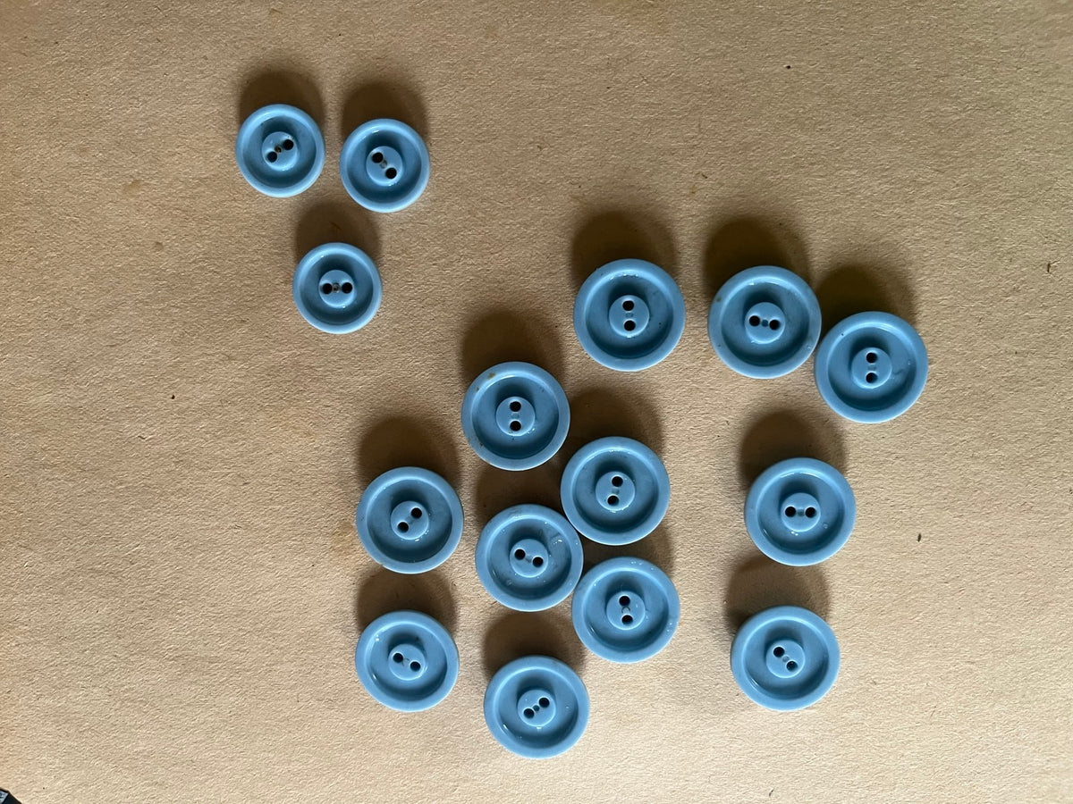 Fifteen powder blue round two-hole buttons in mixed sizes showing recessed backs on brown paper.