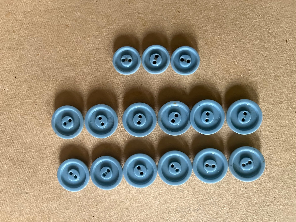 Fifteen powder blue round two-hole buttons in mixed sizes showing recessed backs arranged in rows on brown paper.