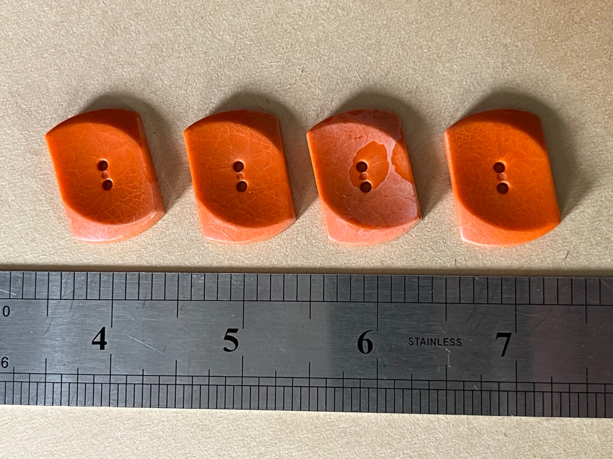 Four rust orange curved rectangular buttons beside a metal ruler on beige paper.