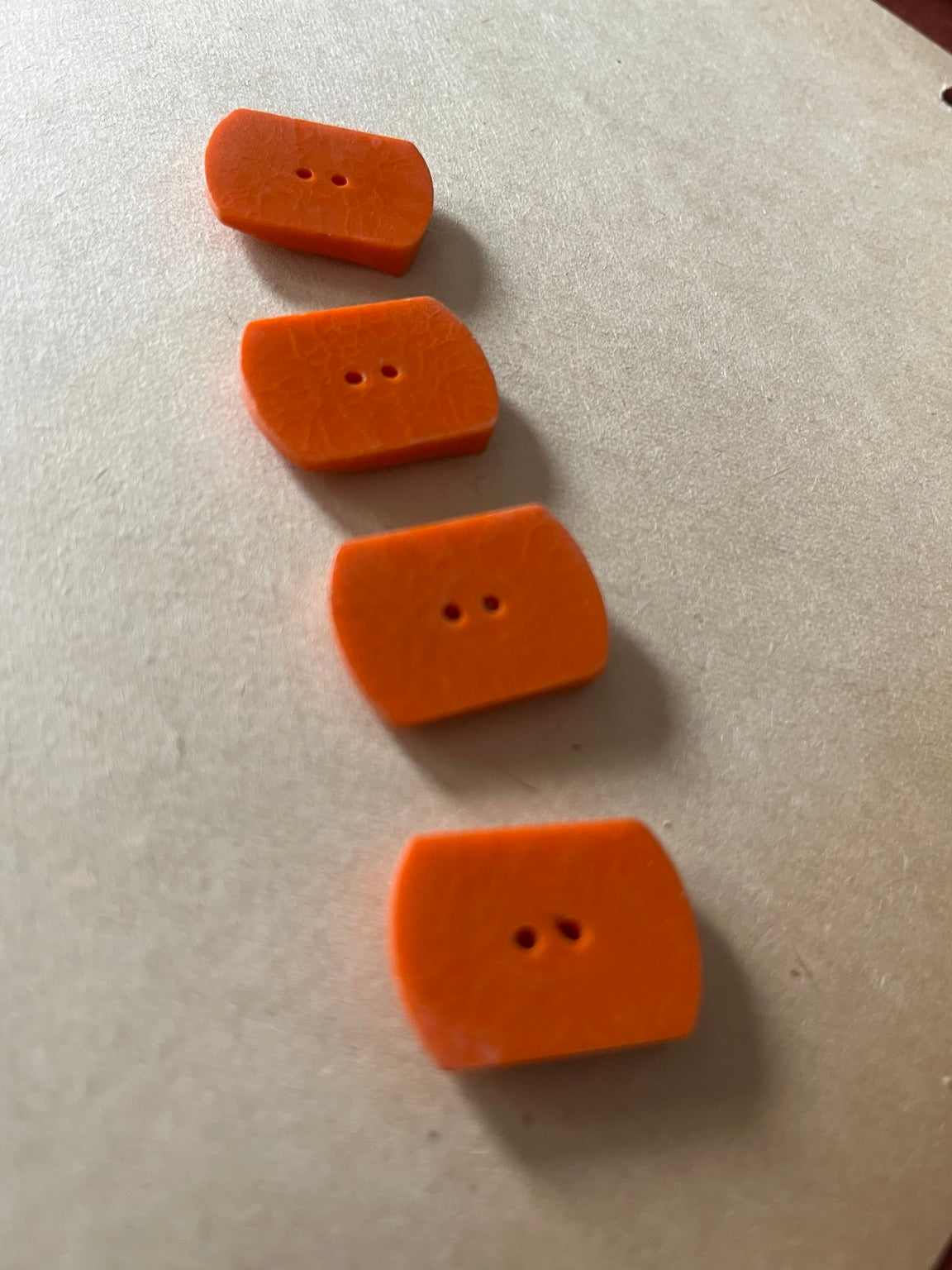 Four rust orange curved rectangular buttons showing two-hole backs on beige paper.