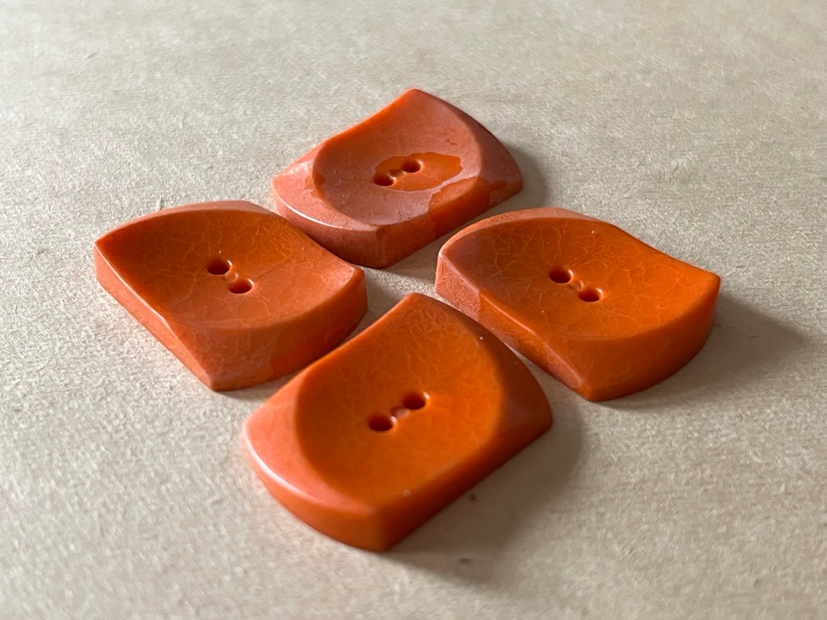 Four rust orange curved rectangular buttons showing recessed two-hole backs on beige paper.