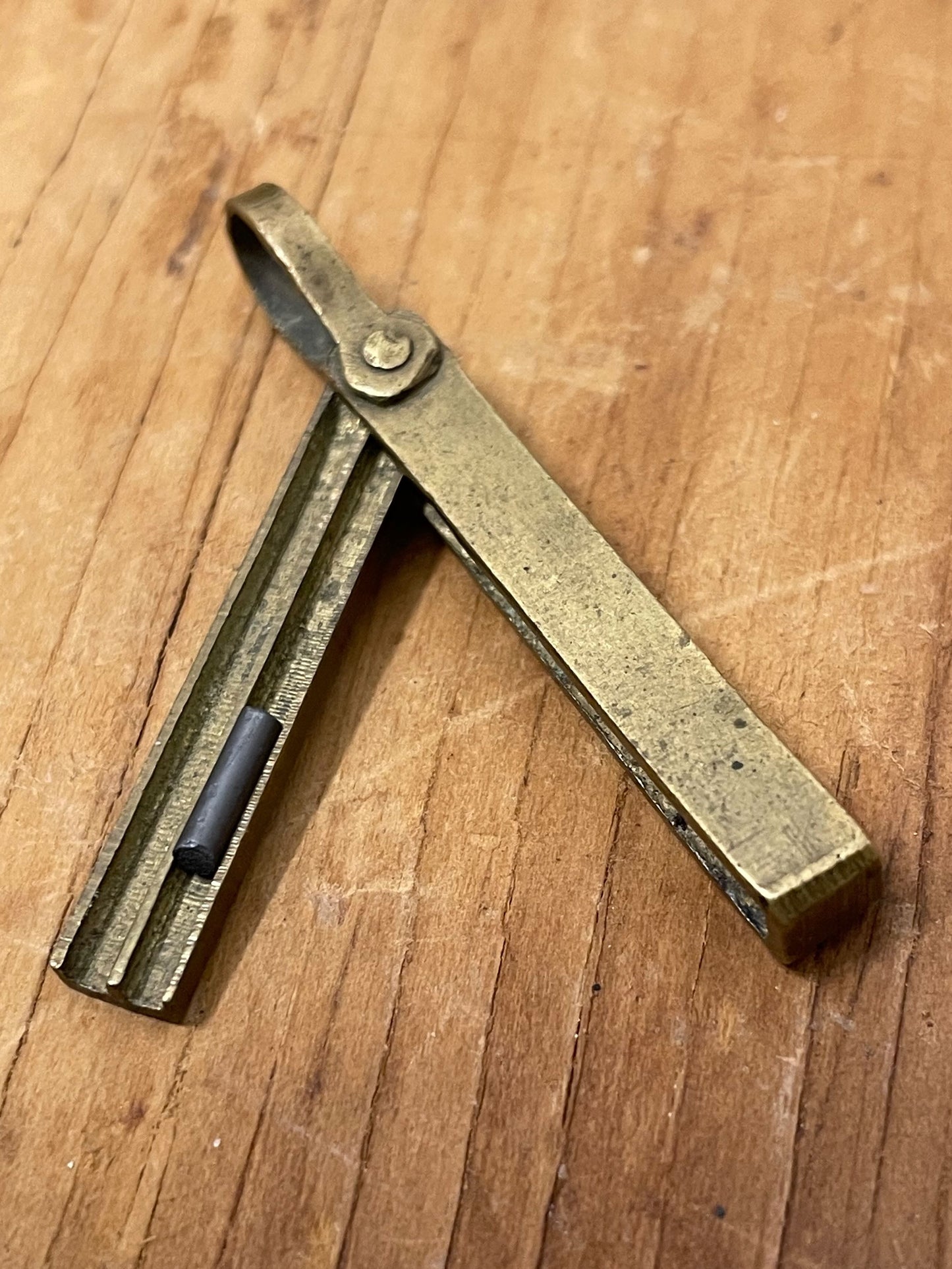Petite brass pendant-style mechanical pencil lead holder on a wooden surface. Small piece of pencil lead pictured for reference.