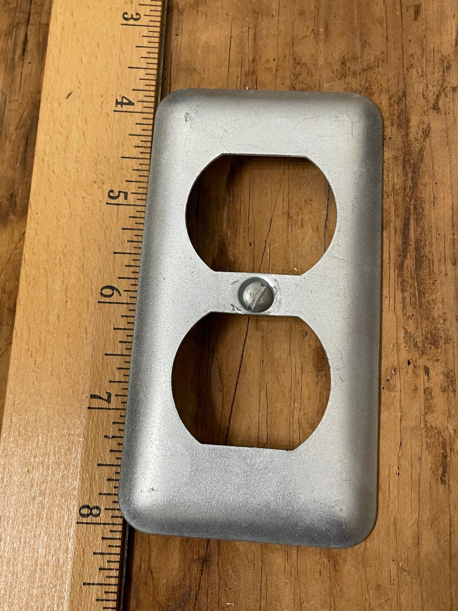 Metal electrical outlet cover on a wooden surface with a ruler for scale.