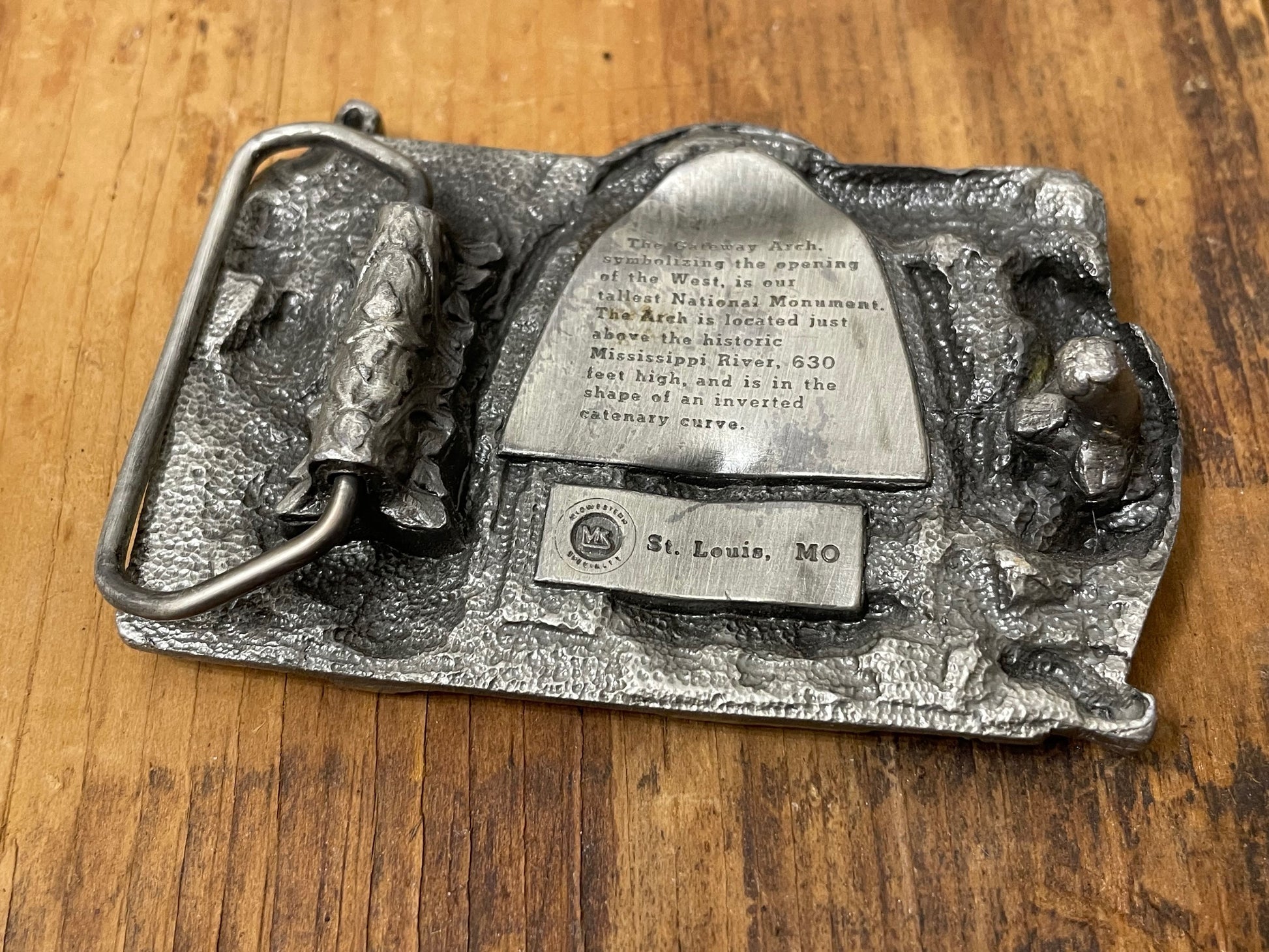 Underside of belt buckle with 'Gateway Saint Louis Arch MO' inscription on a wooden surface