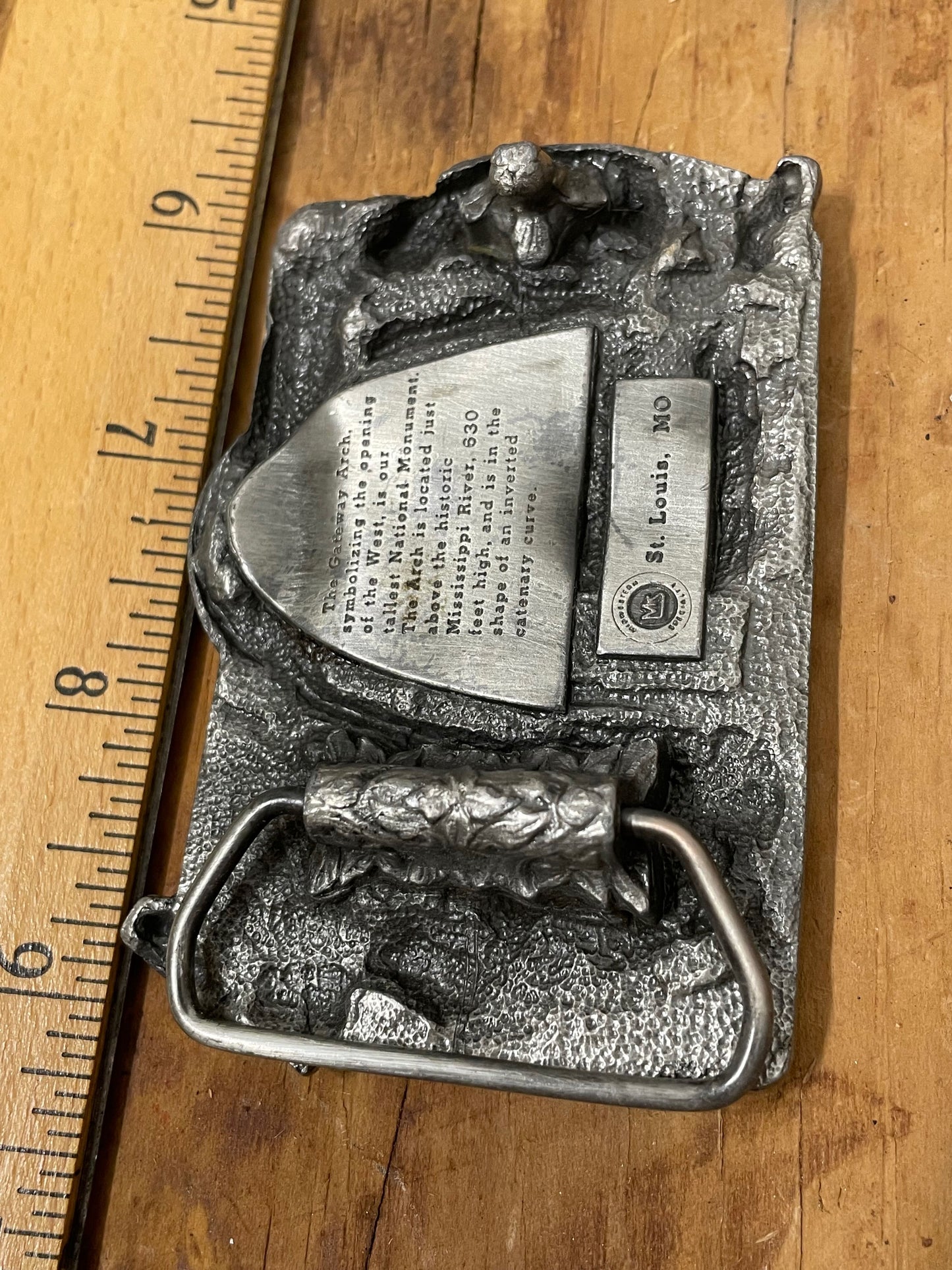 Underside of belt buckle with 'Gateway Saint Louis Arch MO' inscription and a wooden ruler on a wooden surface (buckle measures roughly 3" across).