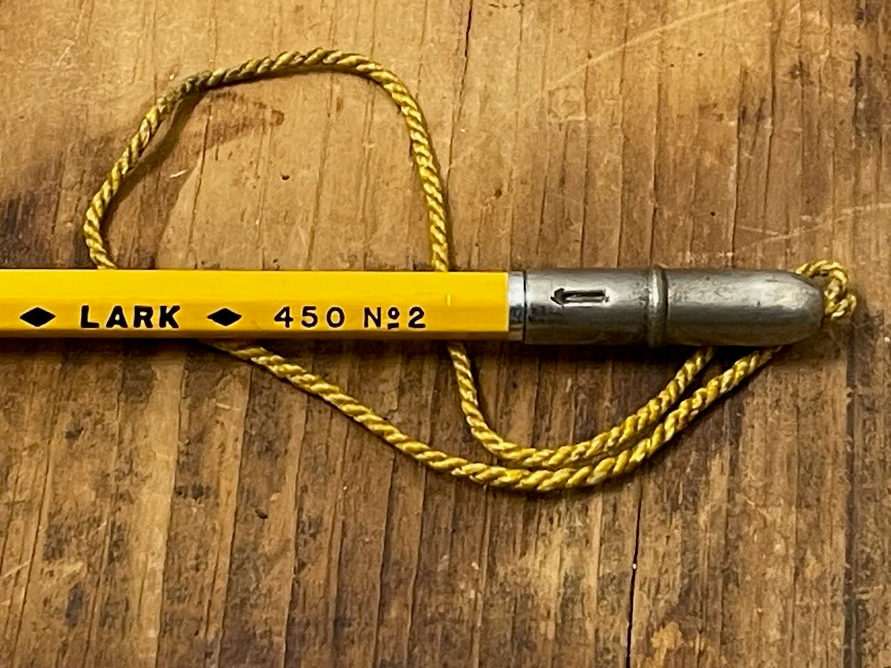Metal pencil topper with attached cord covering the eraser end of a Lark No 2 pencil, on a wooden surface