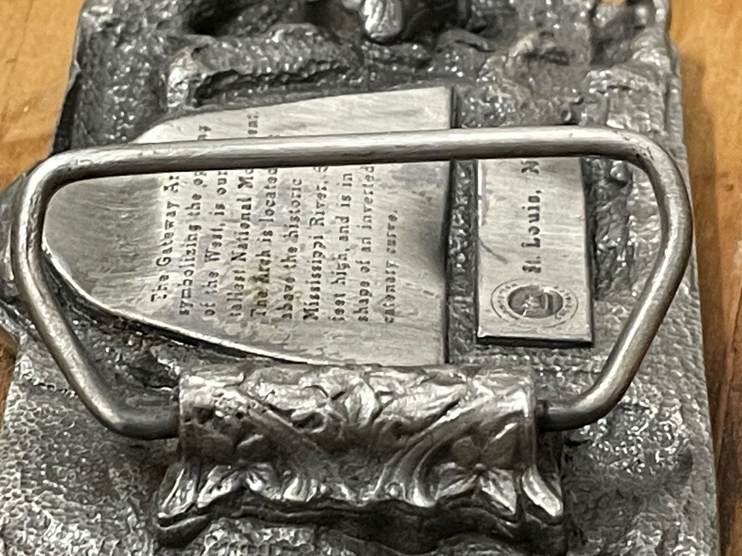 Back closeup of metallic belt buckle with 'Gateway Saint Louis Arch MO' text on a wooden surface