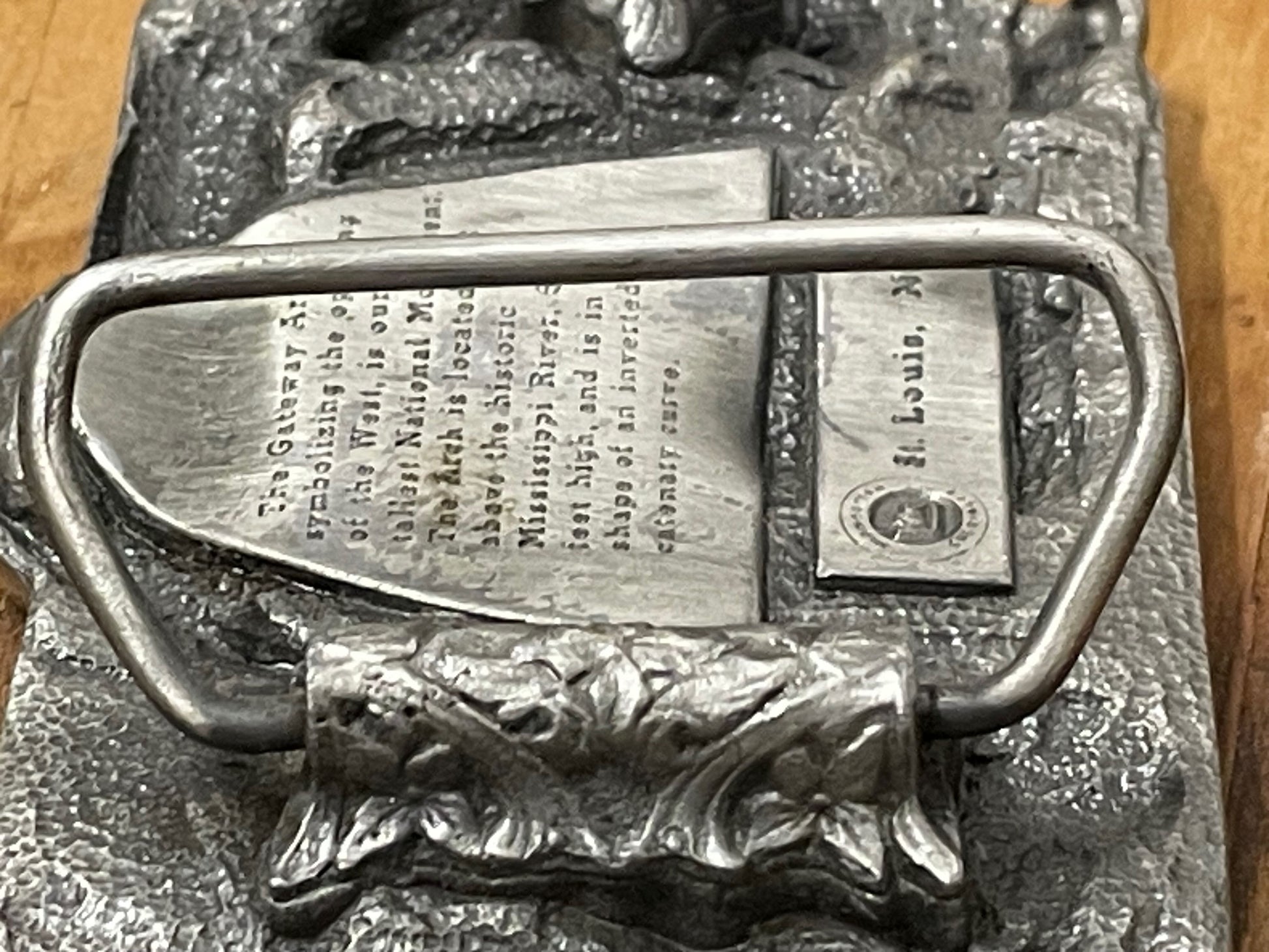 Back closeup of metallic belt buckle with 'Gateway Saint Louis Arch MO' text on a wooden surface