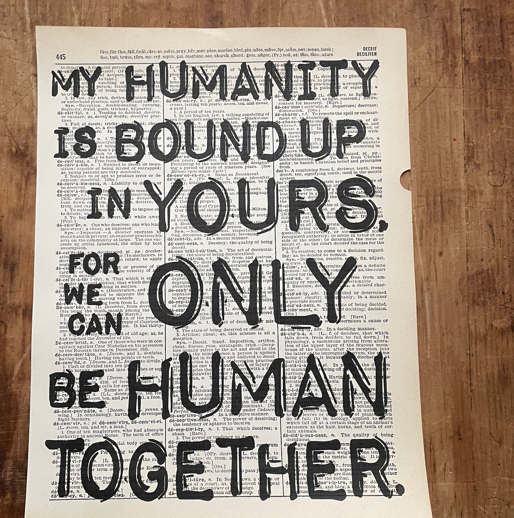 Dictionary page with overprinted motivational quote on a wooden surface:"My humanity is bound up in yours, for we can only be human together."(Desmond Tutu)