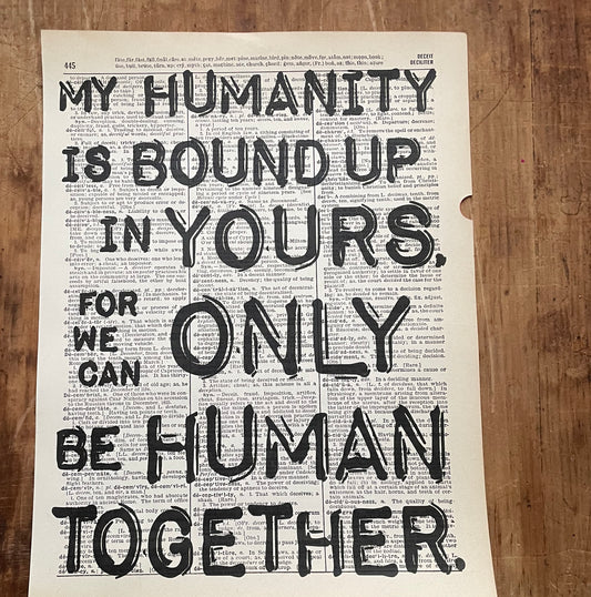Dictionary page with overprinted motivational quote on a wooden surface:"My humanity is bound up in yours, for we can only be human together."(Desmond Tutu)