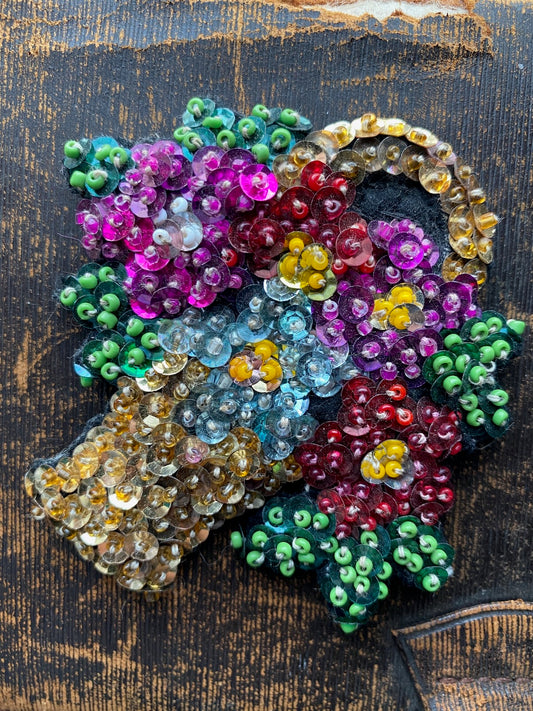 Handmade brooch shaped like a flower basket made using sequins and beads against a distressed black surface.