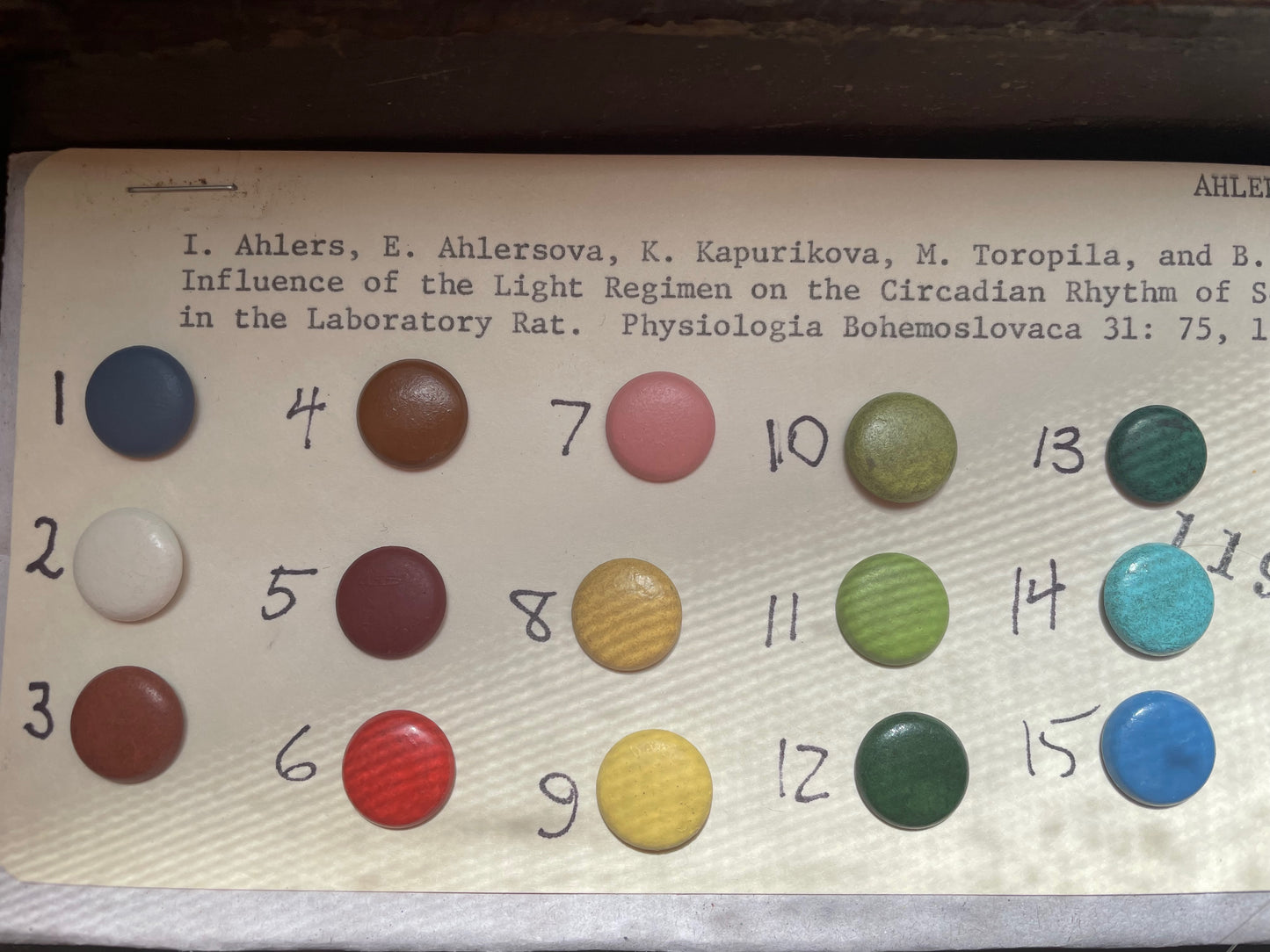 15 numbered, colored decorative upholstery tacks arranged in a grid on a vintage catalog card that described where to find the publication for a scientific experiment about Light on Laboratory Rats' Circadian Rhythms. 