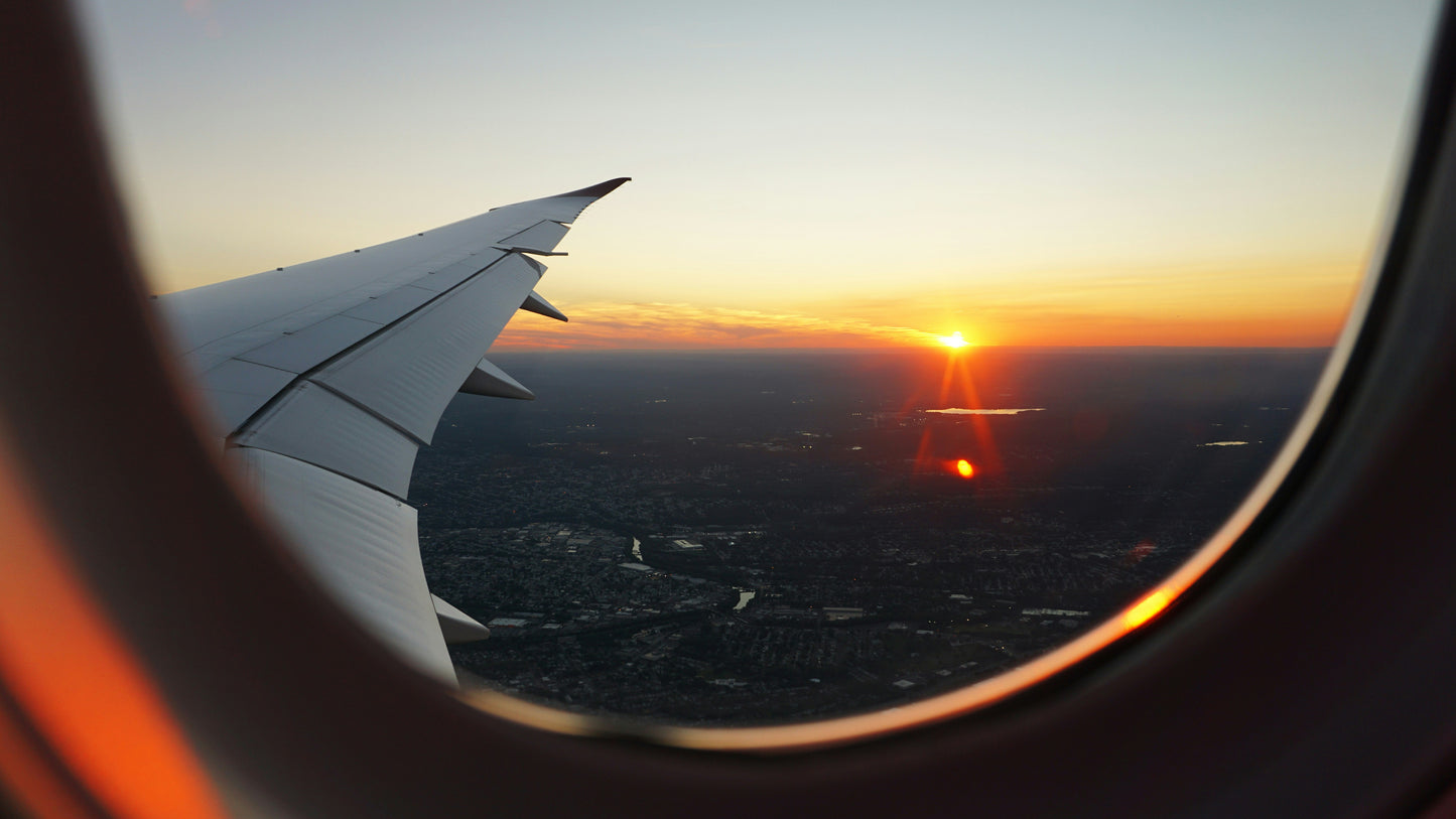 Photo is taken through an airplane window overlooking the wing, a city below, and the sun right at the edge of the horizon. Photo by Eva Darron on Unsplash
